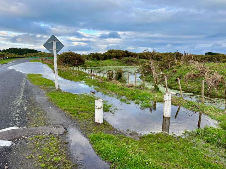 The flooded paddock that has overflowed onto the road, and the grass verge full of water. . 